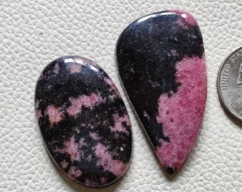 May include: Two cabochon gemstones, one oval and one teardrop shaped, with a black and pink pattern. The stones are polished and have a glossy finish.