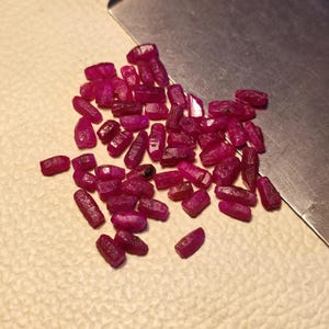 May include: A collection of raw, uncut ruby gemstones. The rubies are a deep, rich red color, with a slightly rough texture. They are scattered on a light-colored surface, with a metal tool in the background. The gemstones vary in size and shape.