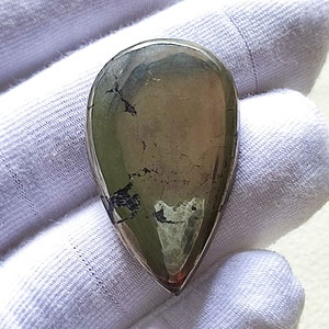 Può includere: Un cabochon di pirite lucido a forma di lacrima, di colore grigio argenteo metallico e con un motivo di venature e inclusiones grigio scuro, quasi nero.