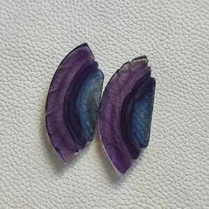 以下が含まれることがあります： 層状の縞模様のある、紫と青の蛍石のcabochonが2つ。cabochonは半円形で、滑らかで磨かれた表面をしています。