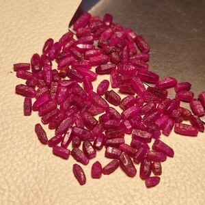 May include: A collection of raw, uncut ruby gemstones. The rubies are a deep, rich red-pink color, with a slightly translucent appearance. They are irregularly shaped, with a rough, natural texture. The gemstones are scattered on a light-colored surface.