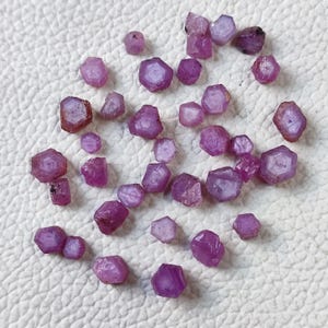 May include: A collection of raw, uncut ruby gemstones in varying sizes. The gemstones display a range of pink and purple hues, with visible crystalline structures. The rough stones are scattered on a white surface.