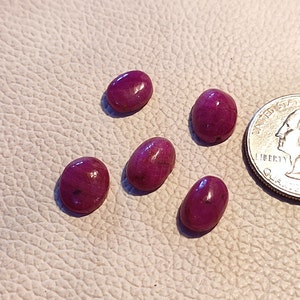 May include: Five oval-shaped, polished ruby gemstones in varying sizes. The gemstones are a deep, rich red color with subtle variations in tone. A US quarter coin is included for size comparison. The gemstones are displayed on a textured, off-white surface.