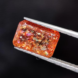 Op de afbeelding: Een rechthoekige, oranje edelsteen met kleurrijke insluitsels, vastgehouden door een zilveren pincet tegen een zwarte achtergrond. Het gepolijste oppervlak is doorschijnend en toont interne details. De edelsteen is ongeveer 1,5 cm lang.