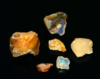 Puede incluir: Una colección de piedras preciosas de ópalo en bruto de varias formas y tamaños. Los ópalos muestran una gama de colores, incluyendo naranja, amarillo y azul, con una textura rugosa y natural. Las piedras preciosas están sobre un fondo negro.