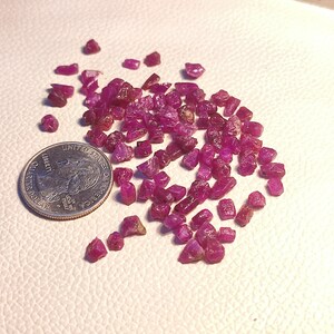 Könnte beinhalten: Eine Sammlung kleiner, rosafarbener Rubinkristalle, einige mit einem rötlicheren Farbton, verstreut auf einer weißen Oberfläche. Eine deutsche 1-Euro-Münze dient als Größenvergleich.