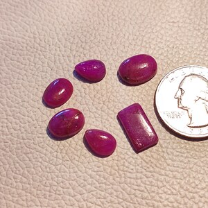 May include: Six pink ruby gemstones of various shapes and sizes. The gemstones are arranged on a white surface. A US quarter is included for scale.