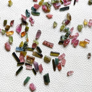 May include: A scattering of small, multi-coloured tourmaline crystals on a white surface. The crystals exhibit a range of colours, such as pink, green, yellow, and blue. The crystals vary in shape and size, some appearing cut or polished.
