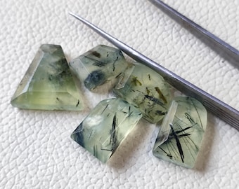 May include: A collection of translucent, light green prehnite gemstones with black inclusions. The gemstones are cut into various shapes, including a triangle and rectangles. A pair of silver tweezers are in the background.