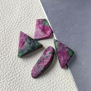 Può includere: Quattro pietre preziose grezze di rubino e zoisite di varie forme. Le pietre preziose mostrano un mix di colori rosa intenso, verde e nero. Le pietre sono poste su una superficie bianca strutturata e un bordo metallico argentato.