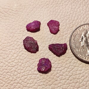 May include: Five small, rough, pink ruby gemstones on a light brown textured surface. A US quarter is included for size comparison.
