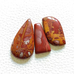 Puede incluir: Tres cabujones de piedra de forma irregular con patrones rojos, naranjas y blancos. Las piedras son lisas y pulidas.