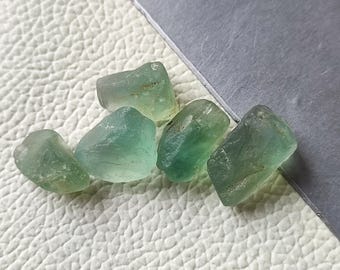 以下が含まれることがあります： 緑と青緑の様々な色合いの、5つの未加工の未切断蛍石原石。石はざらざらした自然な質感で、灰色の対角線の縁がある白い表面に表示されています。