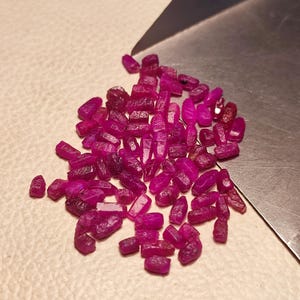 Peut inclure: Un ensemble de pierres précieuses rubis brutes. Les pierres, d'un rouge-rose profond, présentent des formes irrégulières et des tailles variées. Elles sont disposées sur une surface claire, avec un outil métallique en arrière-plan.