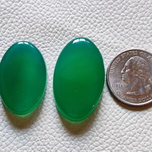 Può includere: Due cabochon ovali in agata verde, uno leggermente più grande dell'altro, accanto a un quarto di dollaro americano per il confronto delle dimensioni.
