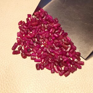 Peut inclure: Un ensemble de pierres précieuses rubis brutes, non taillées, d'un rouge profond. Les pierres sont dispersées sur une surface claire, avec un outil métallique partiellement visible. Les pierres sont probablement mesurées en grammes.