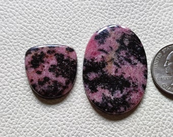 May include: Two polished rhodonite gemstones. One is a rounded rectangle, the other is oval. Both feature a pink base with black veining. A US quarter is next to the oval gemstone for size comparison.