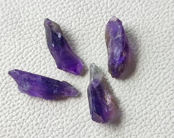 Peut inclure: Quatre pointes de cristaux d'améthyste bruts de tailles variables. Les cristaux présentent une teinte violet foncé avec des zones de couleur plus claire et des imperfections naturelles. Les cristaux sont disposés sur une surface blanche.