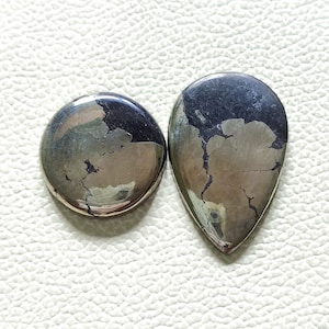 Può includere: Due cabochon di pirite, uno rotondo e uno a forma di lacrima, con un colore argento e nero metallizzato. Le pietre hanno una texture naturale e ruvida.