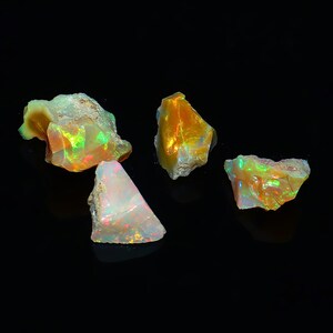 Puede incluir: Cuatro piedras de ópalo natural en bruto con colores iridiscentes de verde, naranja y rojo sobre un fondo negro.