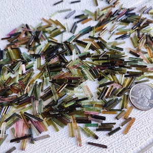 May include: A collection of small, colourful tourmaline crystals in various shades of green, brown, pink, and black. The crystals are arranged on a white surface, with a UK 20 pence coin for scale.
