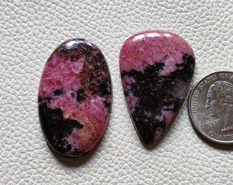 May include: Two pink and black gemstone cabochons, one oval and one teardrop shaped, next to a US quarter for size comparison.