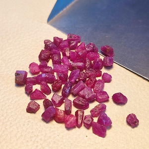 Peut inclure: Un ensemble de pierres précieuses rubis brutes, non taillées, de différentes formes et tailles. Les pierres présentent une couleur rouge intense, avec des nuances plus ou moins translucides. Un outil en métal argenté est en arrière-plan.