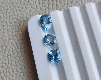 May include: Three light blue gemstones in different cuts: pear, round, and cushion. The gems are displayed on a white, ridged surface, showcasing their clarity and color. The gemstones are likely for jewelry making.