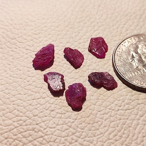 Puede incluir: Seis pequeñas piedras preciosas de rubí rosa sin pulir sobre una superficie blanca texturizada. Se incluye una moneda de veinticinco centavos de dólar estadounidense para la escala.