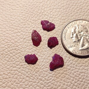 May include: Six small, rough, red ruby gemstones on a light brown leather surface. A US quarter is included for size comparison.