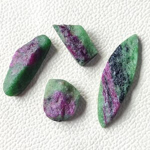 Può includere: Quattro pietre grezze di rubino e zoisite. Le pietre hanno una consistenza ruvida con un mix di colori verde e magenta. Le forme variano da oblunghe a irregolari, mostrando formazioni naturali.