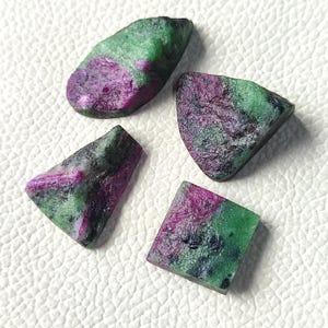 Può includere: Quattro pietre grezze di rubino in zoisite. Le pietre mostrano un mix di colori, tra cui magenta intenso, verde foresta e nero. Le forme variano da rettangolari a triangolari, con una texture naturale e non lucidata. Queste gemme sono ideali per la creazione di gioielli.