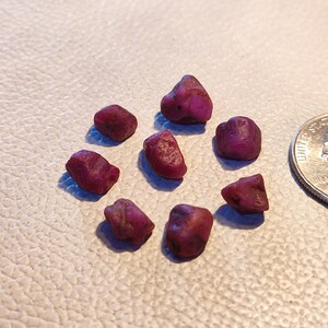 May include: Eight small, rough, purple gemstones, likely rubies, are scattered on a white textured surface. A US quarter is in the background for scale.