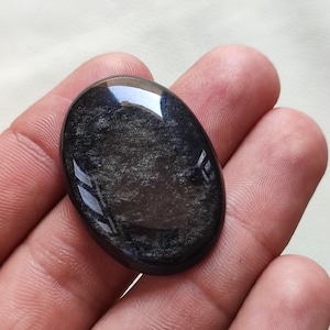 May include: A black oval cabochon gemstone with a shimmering, iridescent finish. The stone has a smooth, polished surface and is set in a bezel setting.