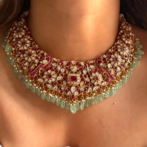 May include: A gold-toned necklace with intricate detailing, featuring red and white gemstones. The necklace has a floral design with a central square-shaped red stone and green teardrop-shaped beads hanging from the bottom.