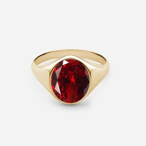May include: A gold signet ring featuring a large, oval-shaped red gemstone.
