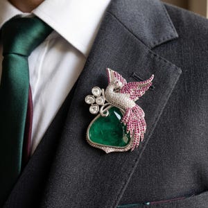 May include: A bird-shaped brooch featuring a large, triangular, deep green gemstone as the body. The bird's head, wings, and tail are encrusted with small, sparkling white and pink stones. A cluster of four round white stones sits near the bird.