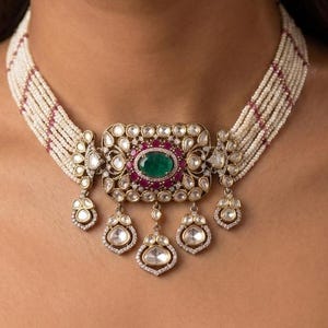 May include: A multi-strand pearl choker necklace featuring a central rectangular pendant with a large oval green stone, surrounded by small clear stones and red accents. The necklace includes five hanging teardrop-shaped charms with clear stones.