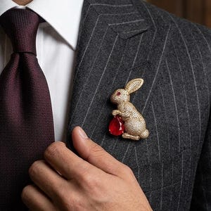 May include: A gold-toned rabbit brooch with a red gemstone, worn on a gray pinstripe suit. The brooch is detailed with small embellishments. The person is also wearing a gold watch and ring, and a burgundy tie.