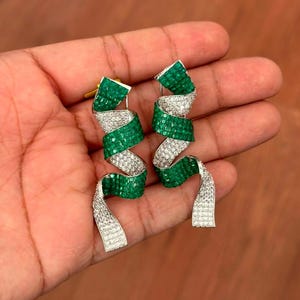 May include: A pair of spiral-shaped dangle earrings featuring alternating bands of pavé-set clear stones and channel-set square-cut green stones. The earrings have a metallic finish and a twisted, ribbon-like design that hangs vertically.