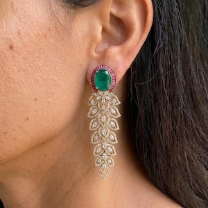 Peut inclure: Boucles d'oreilles dorées élégantes avec une grande émeraude ovale centrale, entourée de petites pierres rouges. Les boucles d'oreilles présentent un motif de feuilles en cascade avec de nombreuses petites pierres claires, créant un look étincelant et luxueux.
