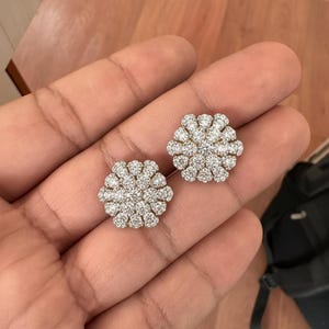 May include: A pair of sparkling, flower-shaped stud earrings. Each earring features a cluster of small, round, clear gemstones, creating a brilliant, floral design. The earrings are set in a silver-colored metal, reflecting light.