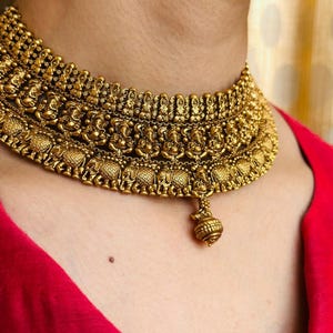 Könnte beinhalten: Ein goldfarbenes Choker-Halsband mit aufwendigen Details. Die Halskette zeigt kleine Figuren und Elefantenmotive mit einem hängenden Anhänger. Der Schmuck wird vor rotem Hintergrund getragen.