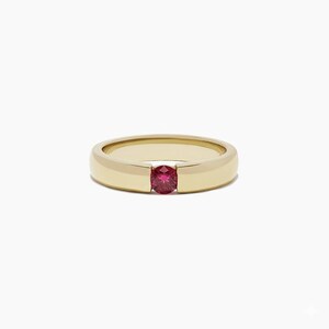 May include: A gold band ring with a round, red gemstone set in the center. The ring has a simple, elegant design with a polished finish. The gemstone is a vibrant red color, adding a pop of color to the ring.