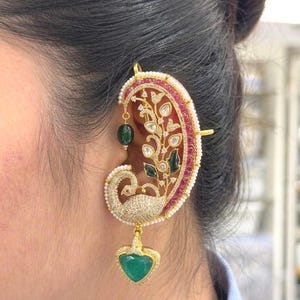 May include: An ornate gold ear cuff featuring a peacock motif encrusted with clear stones, a border of red stones, and small white pearls. The design includes a dangling green heart-shaped stone and a small green bead accenting the upper ear area.