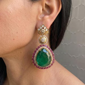 May include: Gold-toned drop earrings with a large, pear-shaped emerald center. The emerald is surrounded by a border of small, red gemstones and a halo of diamonds. The earrings have a floral design at the top.