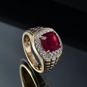 Puede incluir: Un anillo de oro y plata con una gran gema roja oscura cuadrada en el centro, rodeada por un halo de pequeñas gemas claras. La banda del anillo tiene un diseño texturizado.