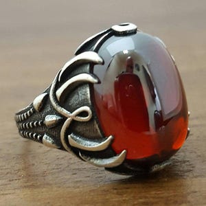 Può includere: Un anello in argento con una grande pietra preziosa rosso scuro, liscia e di forma ovale, incastonata in una fascia di metallo ossidato dettagliata. La lavorazione del metallo presenta intricati motivi in rilievo che ricordano foglie o viticci.