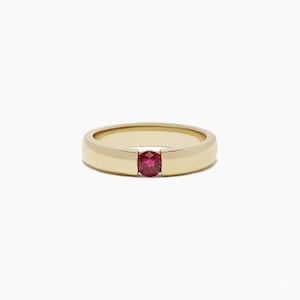 May include: A gold ring featuring a central, round red gemstone. The band has a smooth, polished finish. The ring's design is simple and elegant, with the red gemstone providing a striking contrast to the gold.