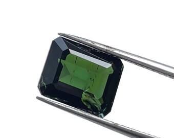 May include: A dark green emerald cut gemstone with a slight black hue, held by tweezers.